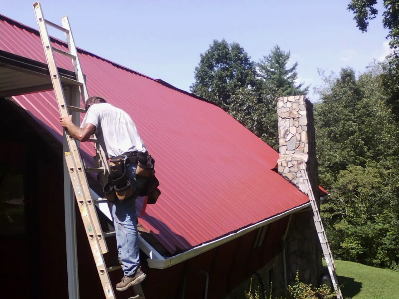 Roofer on a ladder beside a red metal roof and stone chimney — Soffit & Fascia Repair by Roofing Brothers in Blue Mountain